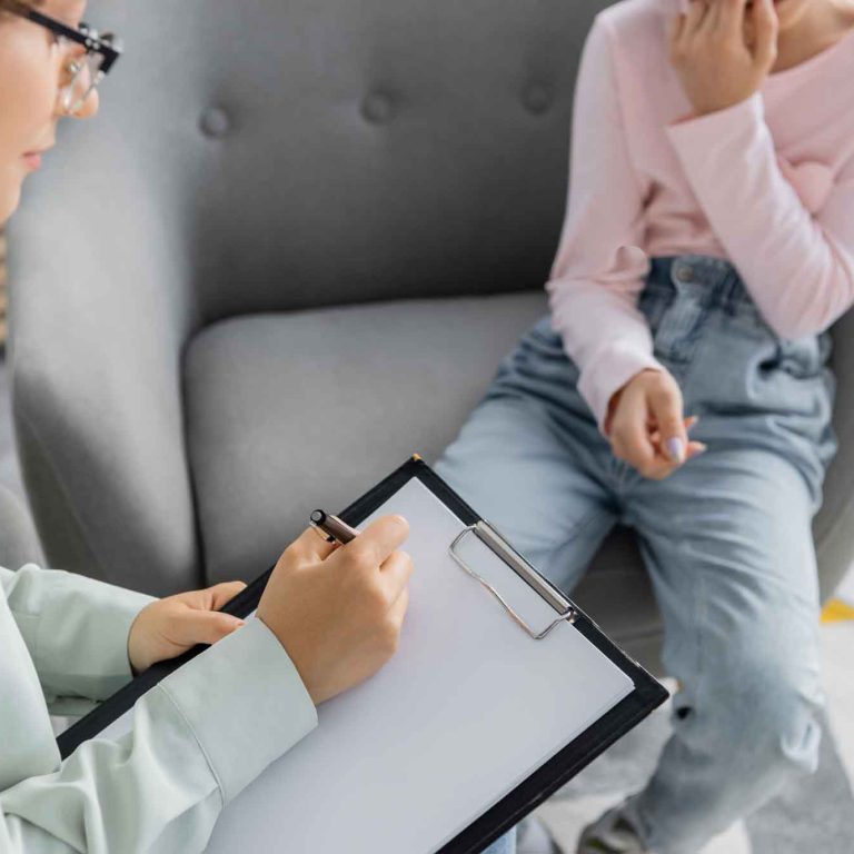 psychologist-writing-clipboard-near-blurred-preteen-patient-consulting-room
